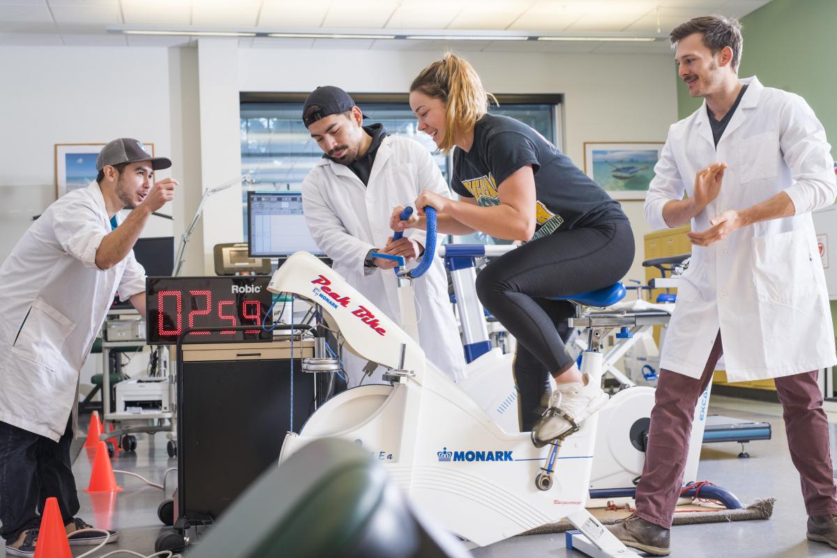 Three students in white lab coats surrounding a fourth student on an exercise bike.